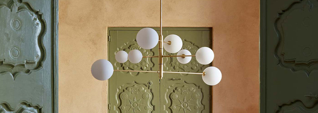 A sculptural brass chandelier with white glass orbs hanging in front of ornate green doors in a warm-toned interior.
