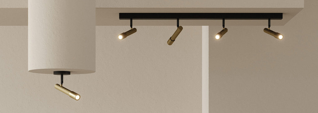 Minimalist ceiling-mounted track lighting with four adjustable brass spotlights and one matching spotlight mounted on an adjacent surface, illuminating a neutral-toned interior.