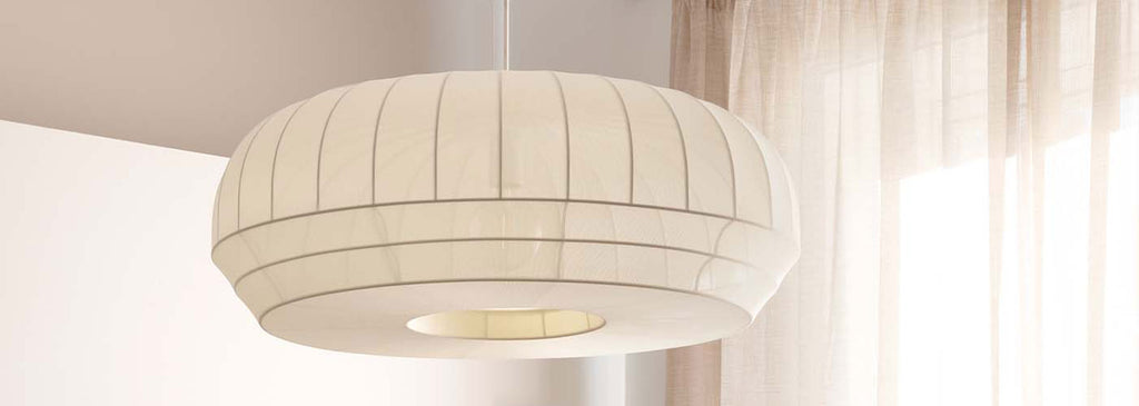 Close-up of the Tradition Pendant Light suspended from a ceiling in a softly lit modern interior. The pendant features a rounded, ribbed design in a warm white finish, diffusing gentle ambient light beside sheer curtains that filter natural daylight.